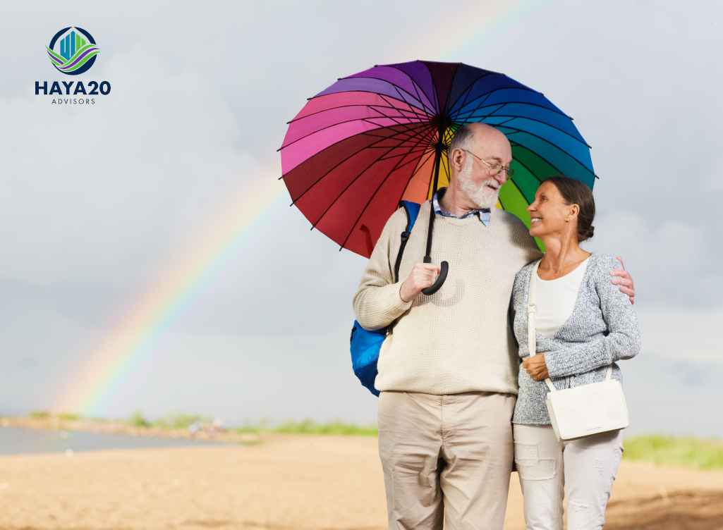 Retired couple standing under a colourful umbrella outdoors with a rainbow in the background.